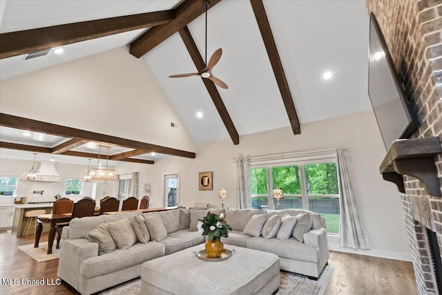 living room featuring ceiling fan with notable chandelier, a brick fireplace, high vaulted ceiling, wood finished floors, and beamed ceiling