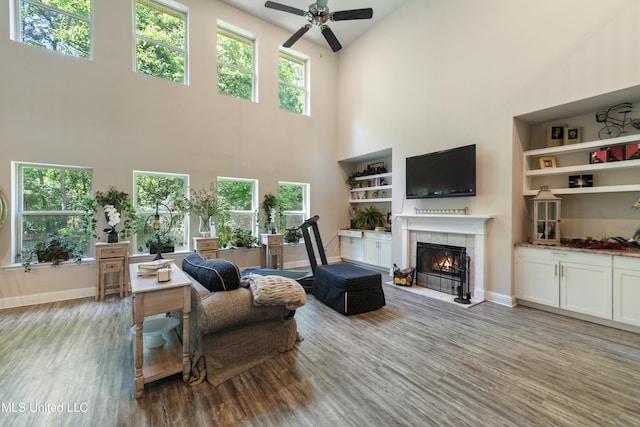 living room with baseboards, a tiled fireplace, ceiling fan, a towering ceiling, and wood finished floors