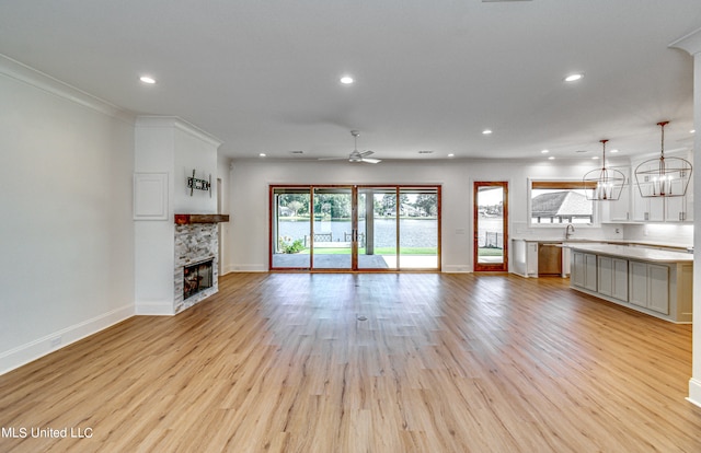 unfurnished living room featuring a stone fireplace, light hardwood / wood-style flooring, sink, crown molding, and ceiling fan with notable chandelier