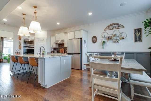 kitchen with under cabinet range hood, ornamental molding, stainless steel appliances, white cabinetry, and dark wood-style flooring