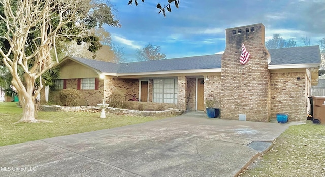 ranch-style home featuring brick siding, a shingled roof, driveway, a chimney, and a front yard