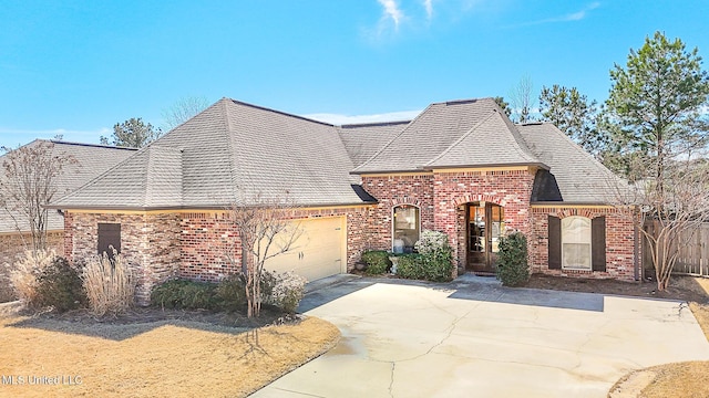 french country home with brick siding, driveway, an attached garage, and roof with shingles