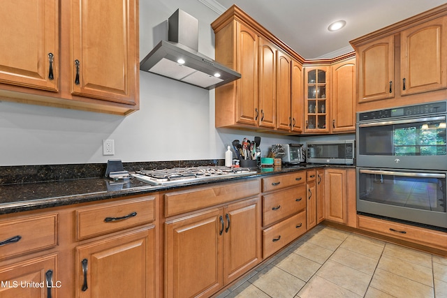 kitchen featuring wall chimney exhaust hood, dark stone countertops, crown molding, light tile patterned flooring, and appliances with stainless steel finishes