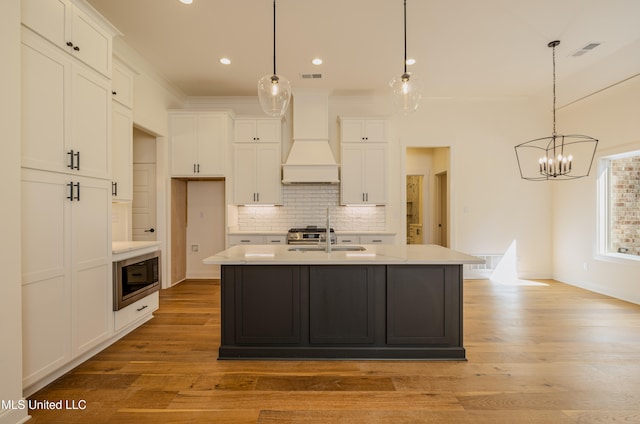 kitchen featuring premium range hood, a kitchen island with sink, stainless steel microwave, and pendant lighting