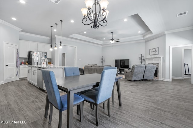 dining area featuring visible vents, ceiling fan with notable chandelier, a tray ceiling, wood finished floors, and a premium fireplace