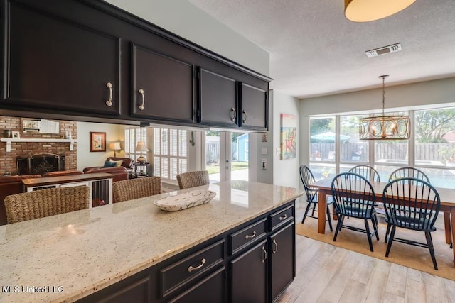 kitchen with light wood-style floors, light stone counters, a brick fireplace, decorative light fixtures, and dark cabinets