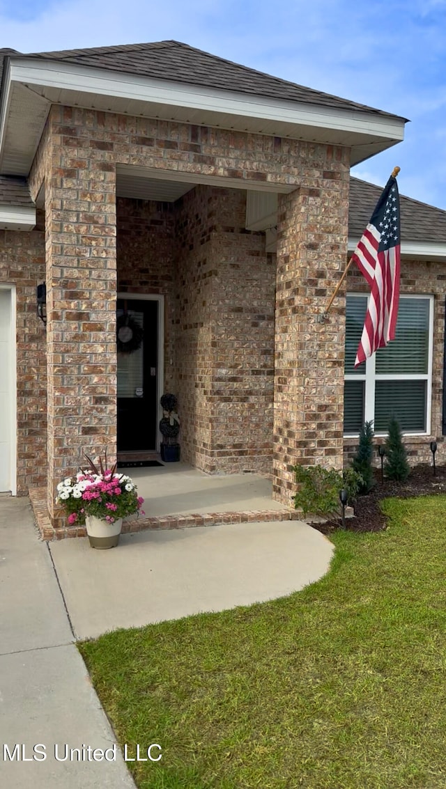 view of exterior entry featuring a yard, brick siding, and a shingled roof