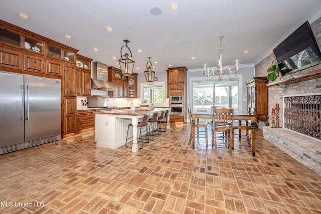 kitchen with brown cabinetry, wall chimney exhaust hood, built in appliances, and brick floor