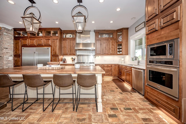 kitchen with wall chimney range hood, decorative backsplash, brown cabinets, a sink, and built in appliances