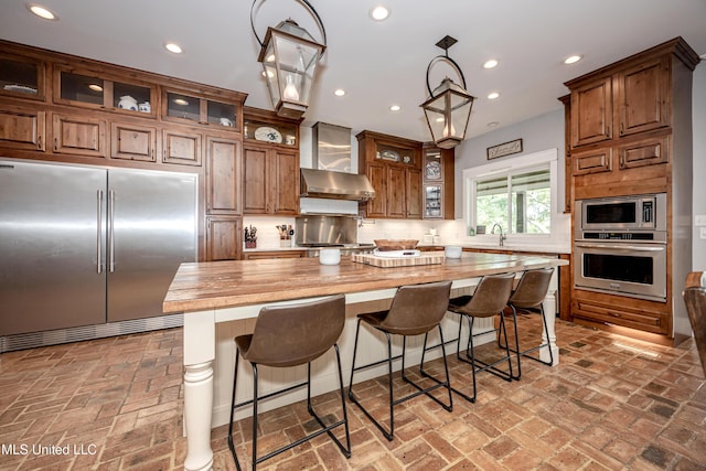 kitchen with wall chimney range hood, built in appliances, brick floor, and butcher block countertops