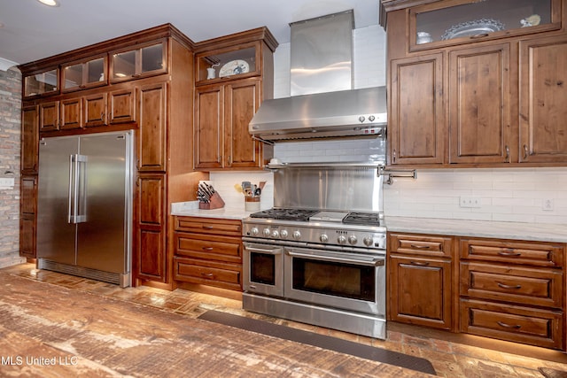 kitchen featuring premium appliances, decorative backsplash, brown cabinetry, and wall chimney exhaust hood