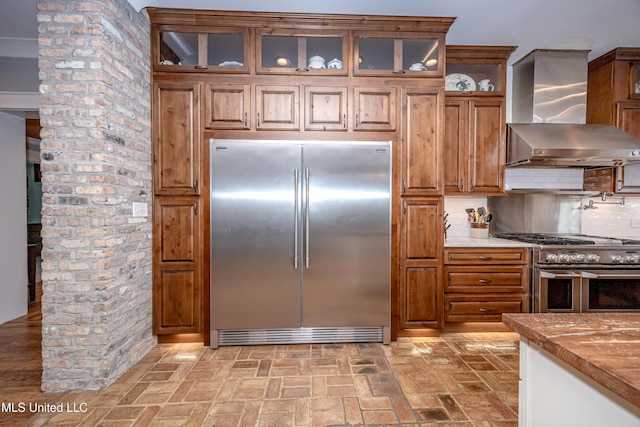 kitchen with wall chimney range hood, brick patterned floor, brown cabinetry, stainless steel appliances, and tasteful backsplash