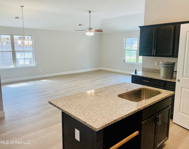 kitchen featuring light wood-type flooring, ceiling fan with notable chandelier, pendant lighting, light stone counters, and a kitchen island with sink