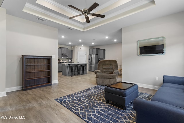living room featuring ceiling fan, visible vents, a raised ceiling, light wood finished floors, and ornamental molding