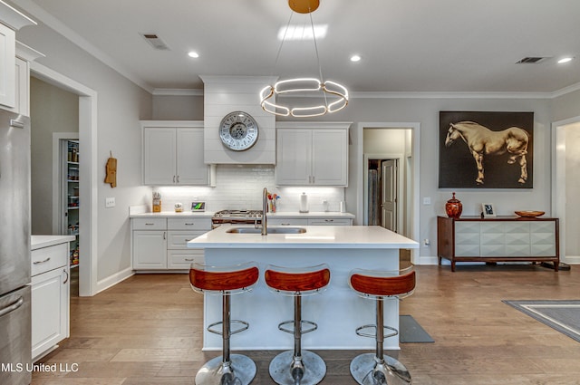 kitchen featuring sink, stainless steel refrigerator, white cabinets, a center island with sink, and decorative light fixtures