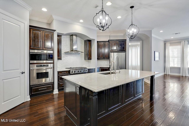 kitchen featuring dark wood-type flooring, stainless steel appliances, wall chimney range hood, pendant lighting, and a center island with sink