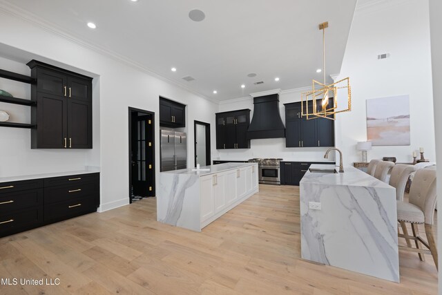 kitchen with light wood finished floors, high end appliances, a kitchen island with sink, dark cabinetry, and premium range hood