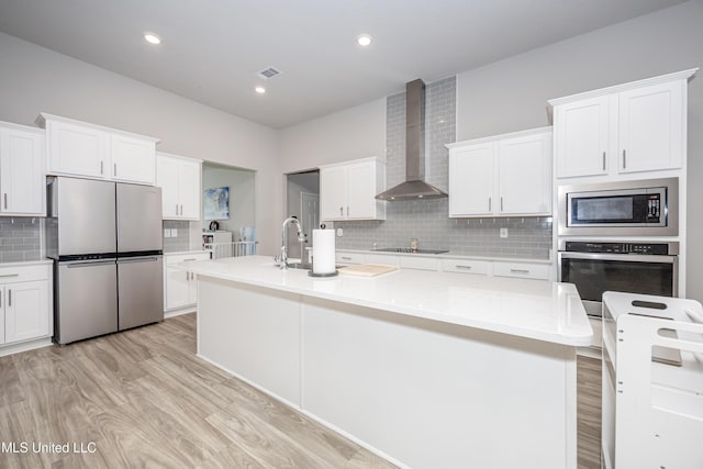 kitchen with light wood-style flooring, appliances with stainless steel finishes, wall chimney exhaust hood, a kitchen island with sink, and backsplash