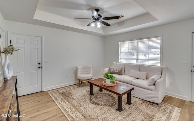 living area featuring baseboards, a raised ceiling, a ceiling fan, and light wood-style floors