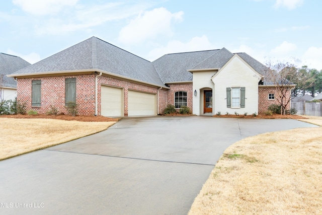 french provincial home featuring a shingled roof, brick siding, driveway, and a garage