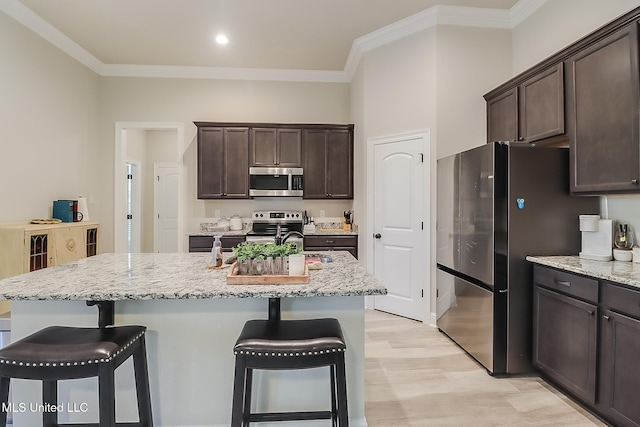 kitchen with light stone countertops, ornamental molding, an island with sink, dark brown cabinetry, and stainless steel appliances