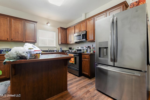 kitchen featuring stainless steel appliances, dark countertops, brown cabinetry, a sink, and wood finished floors