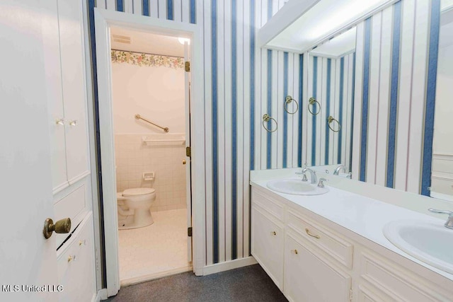 bathroom featuring double vanity, a sink, toilet, and wallpapered walls