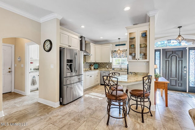 kitchen with wall chimney range hood, stainless steel appliances, decorative light fixtures, light stone counters, and ornamental molding