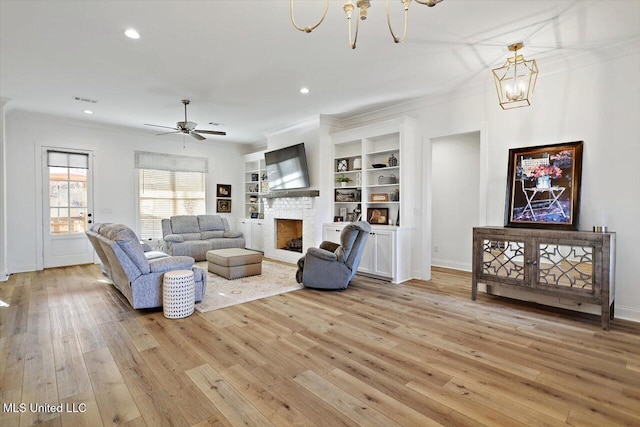 living room with light hardwood / wood-style floors, crown molding, ceiling fan with notable chandelier, built in features, and a stone fireplace