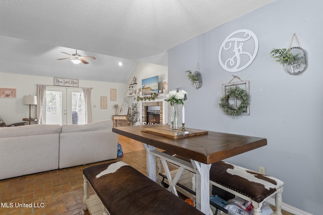 dining space with lofted ceiling, ceiling fan, a textured ceiling, a brick fireplace, and french doors