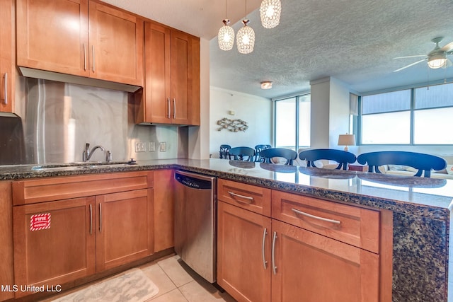 kitchen with light tile patterned floors, a sink, stainless steel dishwasher, brown cabinets, and dark stone countertops