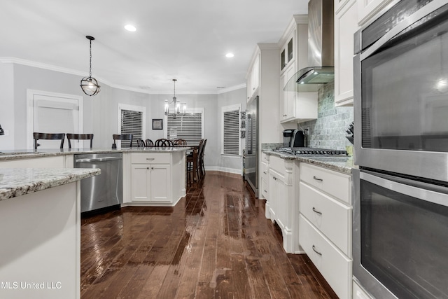 kitchen featuring appliances with stainless steel finishes, wall chimney exhaust hood, ornamental molding, white cabinets, and decorative light fixtures