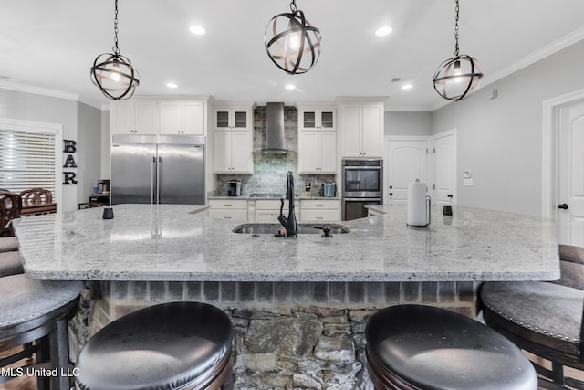 kitchen featuring stainless steel appliances, wall chimney range hood, a sink, crown molding, and a large island with sink