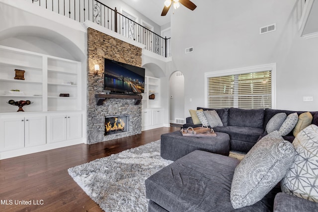 living room featuring built in features, ceiling fan, a towering ceiling, a stone fireplace, and arched walkways