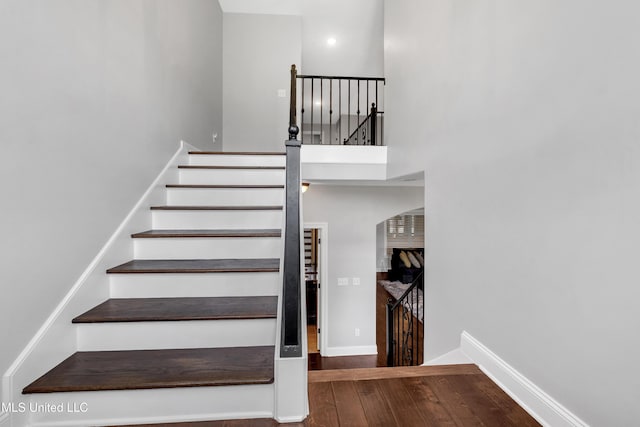 staircase featuring wood finished floors, a towering ceiling, arched walkways, and baseboards