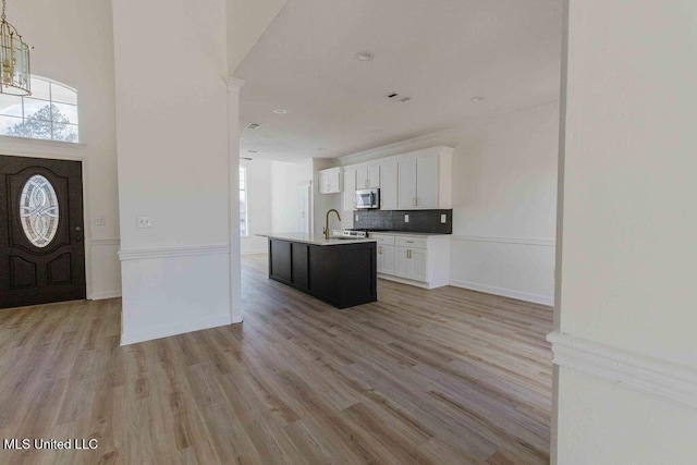 kitchen with a sink, white cabinetry, decorative backsplash, stainless steel microwave, and a center island with sink