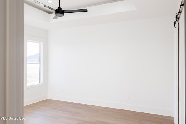 empty room with a barn door, a tray ceiling, ceiling fan, and light hardwood / wood-style floors