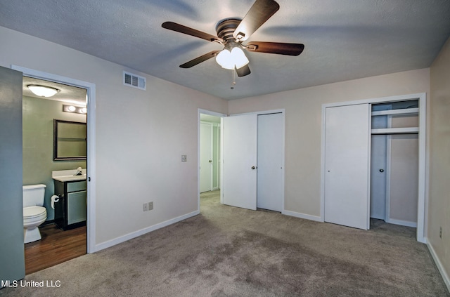 unfurnished bedroom featuring ceiling fan, a textured ceiling, multiple closets, ensuite bathroom, and light colored carpet