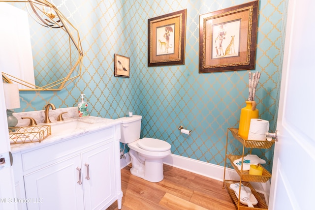 half bathroom with toilet, wood finished floors, vanity, baseboards, and wallpapered walls