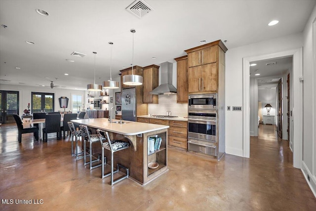 kitchen with appliances with stainless steel finishes, backsplash, wall chimney exhaust hood, pendant lighting, and a center island with sink