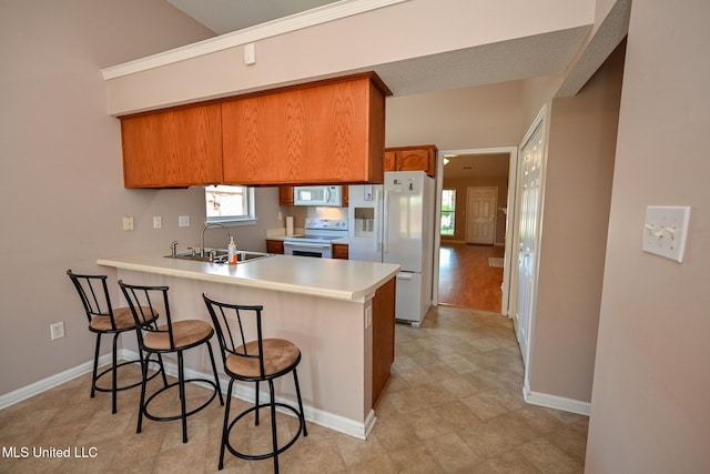 kitchen featuring a sink, a peninsula, brown cabinetry, white appliances, and a kitchen breakfast bar