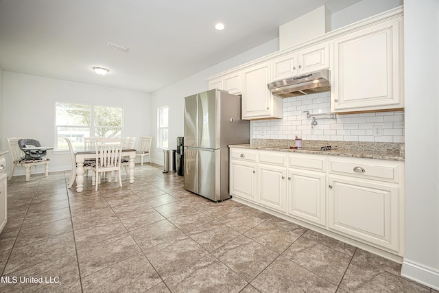 kitchen featuring baseboards, under cabinet range hood, light stone counters, decorative backsplash, and freestanding refrigerator