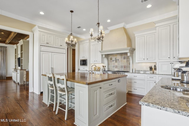 kitchen featuring premium range hood, dark wood-style flooring, a kitchen island with sink, built in appliances, and white cabinetry