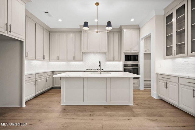 kitchen featuring a kitchen island with sink, appliances with stainless steel finishes, glass insert cabinets, and light countertops