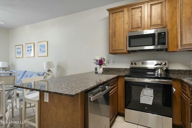 kitchen featuring brown cabinets, stainless steel appliances, dark stone counters, a peninsula, and light tile patterned floors