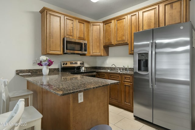 kitchen featuring brown cabinets, appliances with stainless steel finishes, a peninsula, dark stone countertops, and light tile patterned flooring