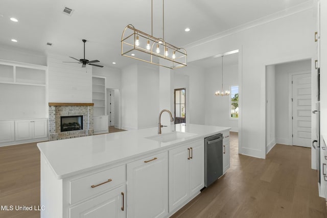 kitchen with ceiling fan with notable chandelier, stainless steel dishwasher, wood-type flooring, a fireplace, and a center island with sink