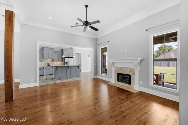 unfurnished living room with crown molding, a sink, a tiled fireplace, light wood-style flooring, and a ceiling fan