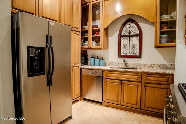 kitchen featuring brown cabinets, appliances with stainless steel finishes, glass insert cabinets, a sink, and light stone countertops