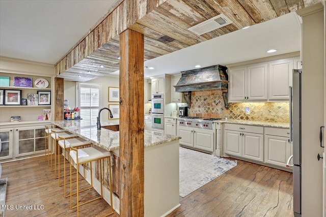 kitchen featuring custom exhaust hood, light hardwood / wood-style flooring, stainless steel appliances, light stone countertops, and backsplash